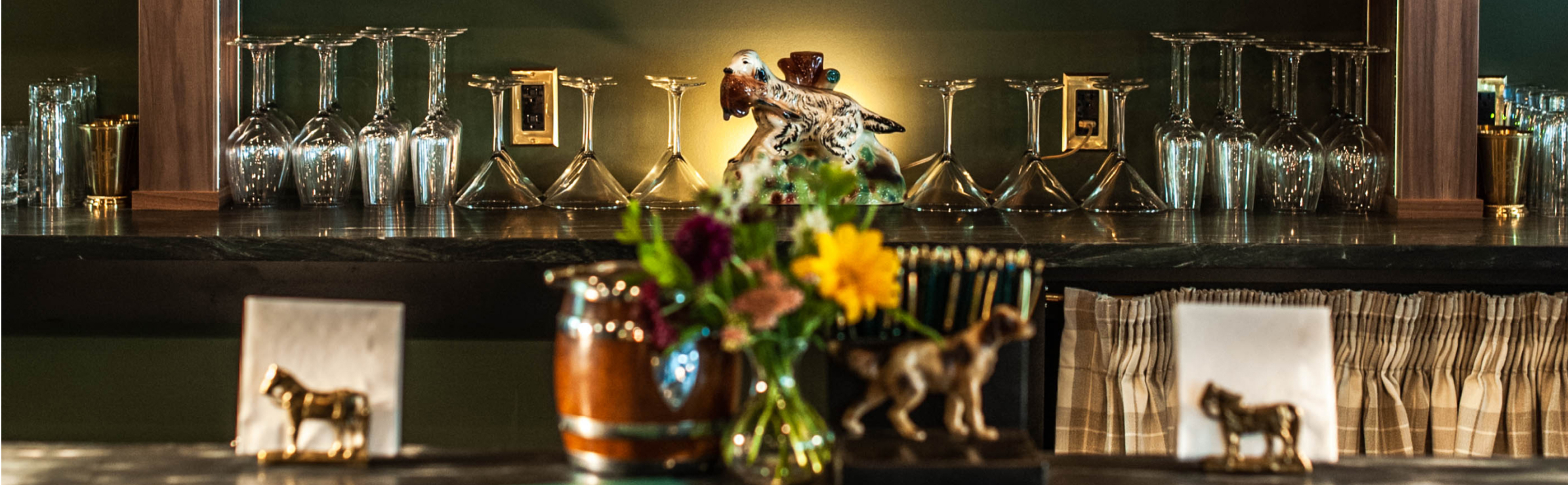 Bar back featuring neatly arranged bar glasses of different shapes. Decor includes vase of flowers and hunting dogs.