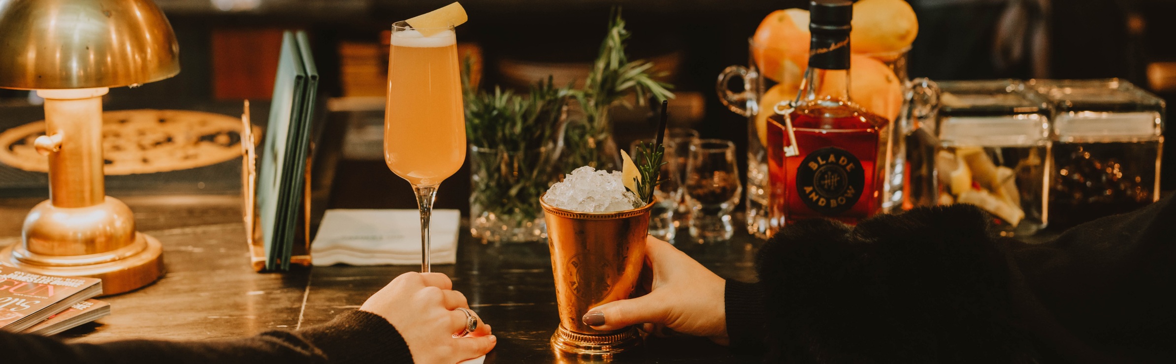 Two different Blade and Bow cocktails being held by two different hands on a bar counter. In the background, a bottle of Blade and Bow whiskey and cocktail garnishes.