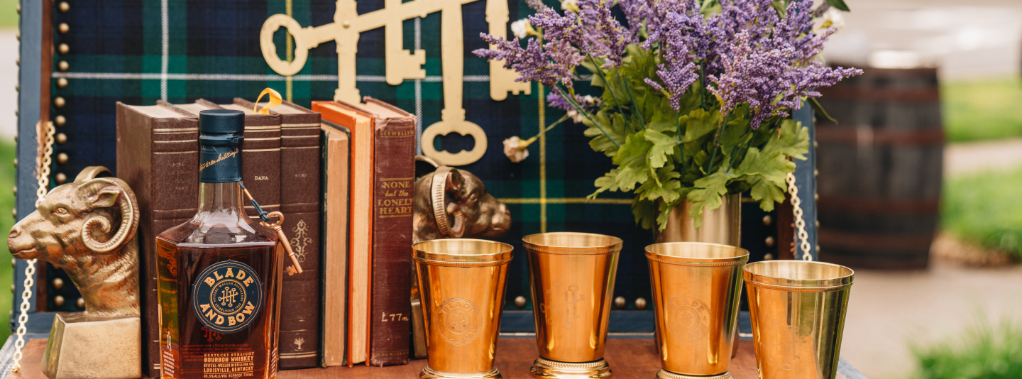 Two glasses on a table with a bottle of Blade and Bow whiskey; potted flowers in background.