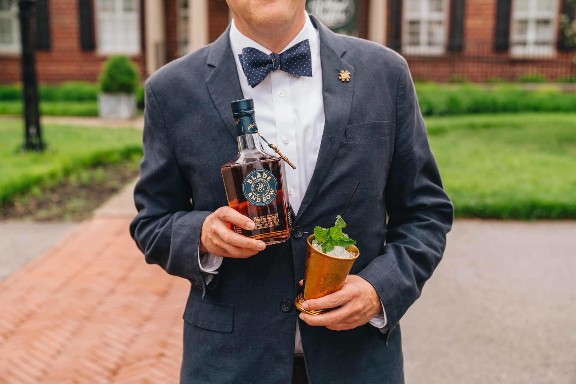 Man in a tuxedo holding a bottle of Blade and Bow whiskey and a brass cup. Out of focus in the background, a dog faces forward.